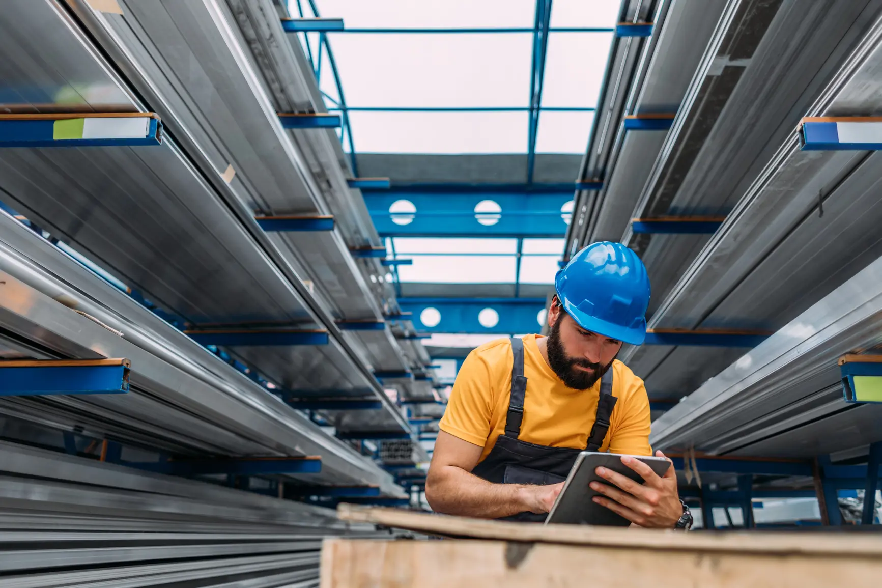 A warehouse worker in a hard hat is engaged with a tablet, likely managing incoming B2B ecommerce orders, surrounded by warehouse shelves filled with goods.